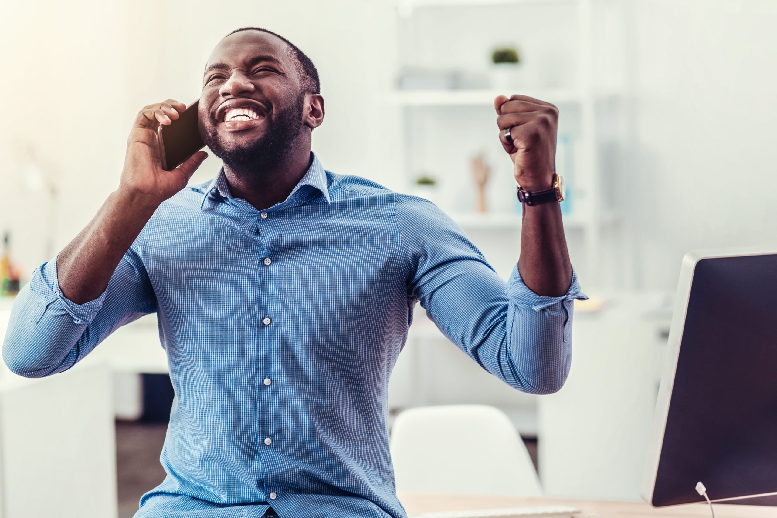 Yay. Waist up shot of a radiant young man cannot keep his emotions inside and triumphing with his hands raised while having a phone conversation at work.