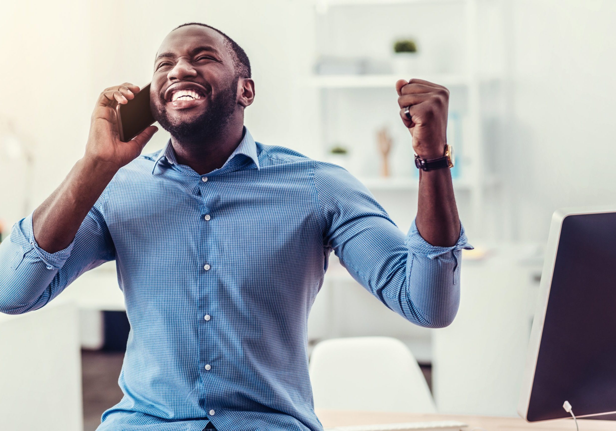 Yay. Waist up shot of a radiant young man cannot keep his emotions inside and triumphing with his hands raised while having a phone conversation at work.